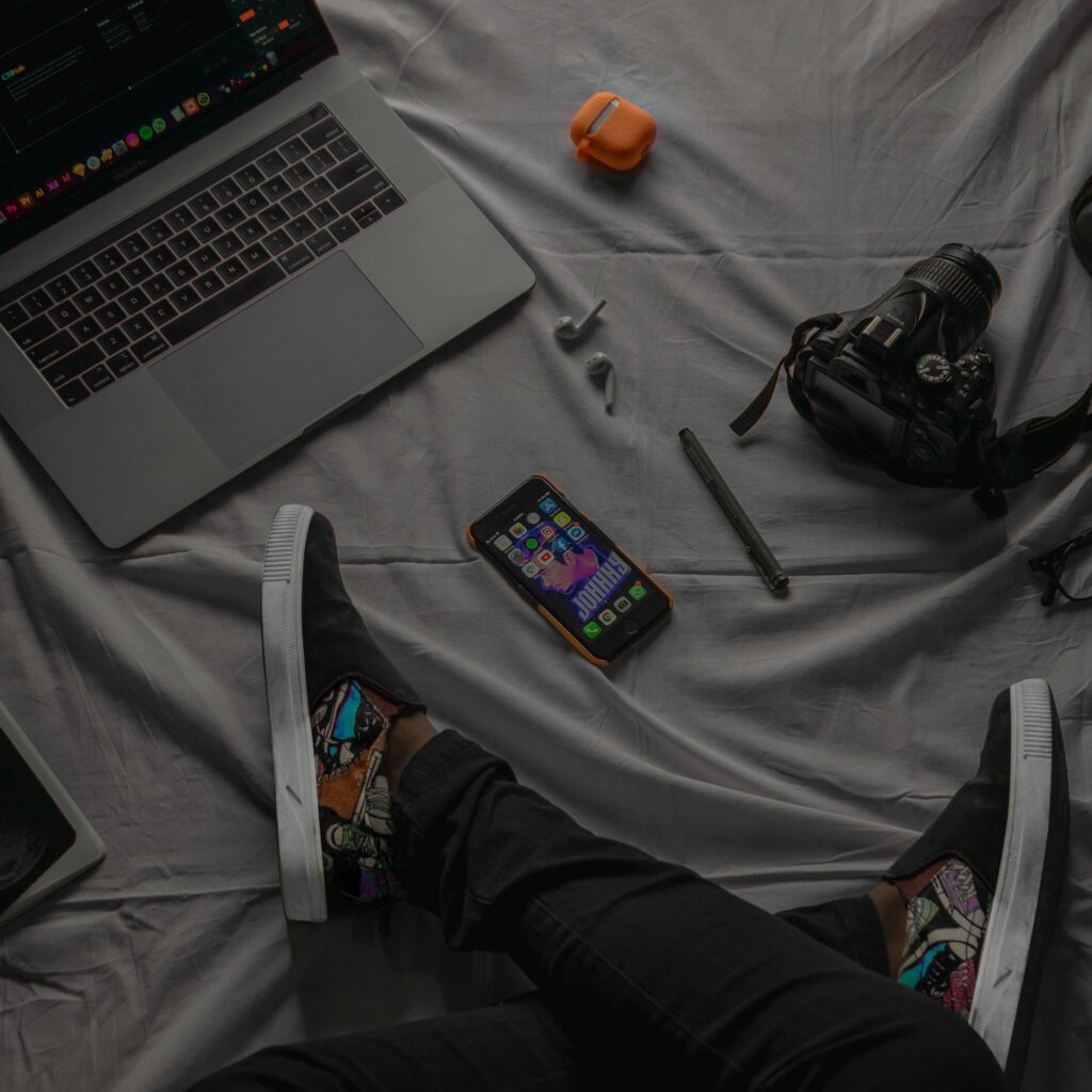 Flat lay of modern tech gadgets on a gray blanket, featuring laptop, camera, and sneakers.